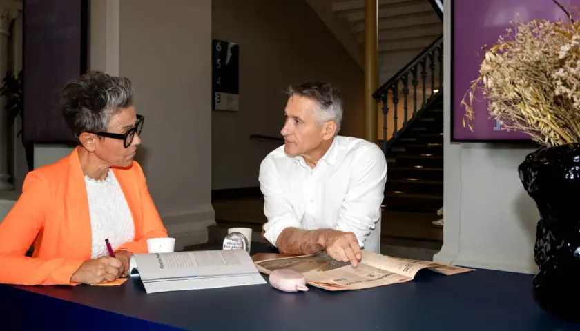 Två personer diskuterar vid ett bord, en kvinna med orange kavaj antecknar, en man pekar på en tidning. Kanske ett möte om administrativa jobb.