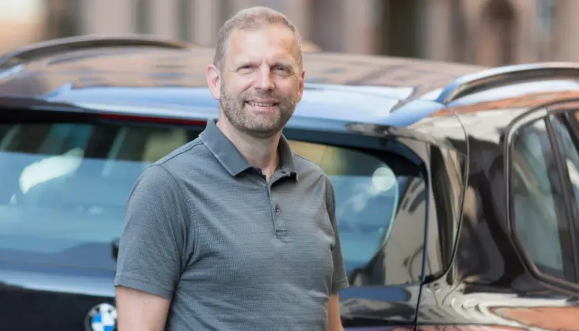 En glad man står bredvid en bil. Kanske funderar han på att leasa en begagnad bil för en bekväm resa.