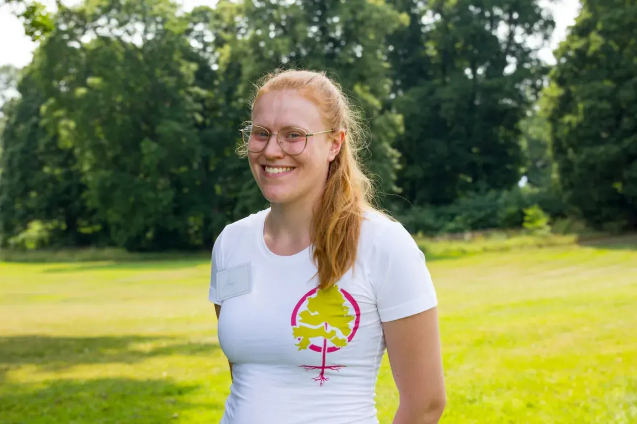 Glad kvinna med glasögon och rött hår ler i en vit t-shirt med ett trädmotiv. Hon verkar trivas med att jobba i naturen.