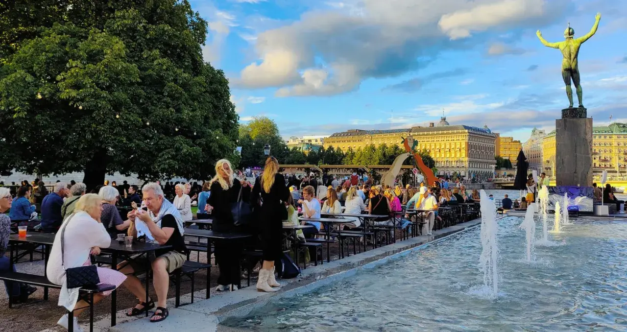 Folk njuter av en sommarkväll med mat och dryck vid fontänen, kanske efter en gratis konsert i Stockholm.