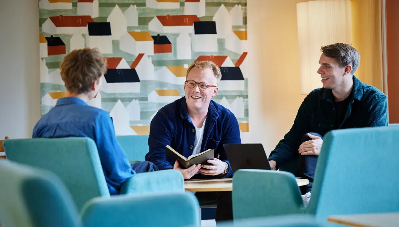 Tre män diskuterar vid ett bord, en med en öppen bok. Kanske en del av deras teologi utbildning.
