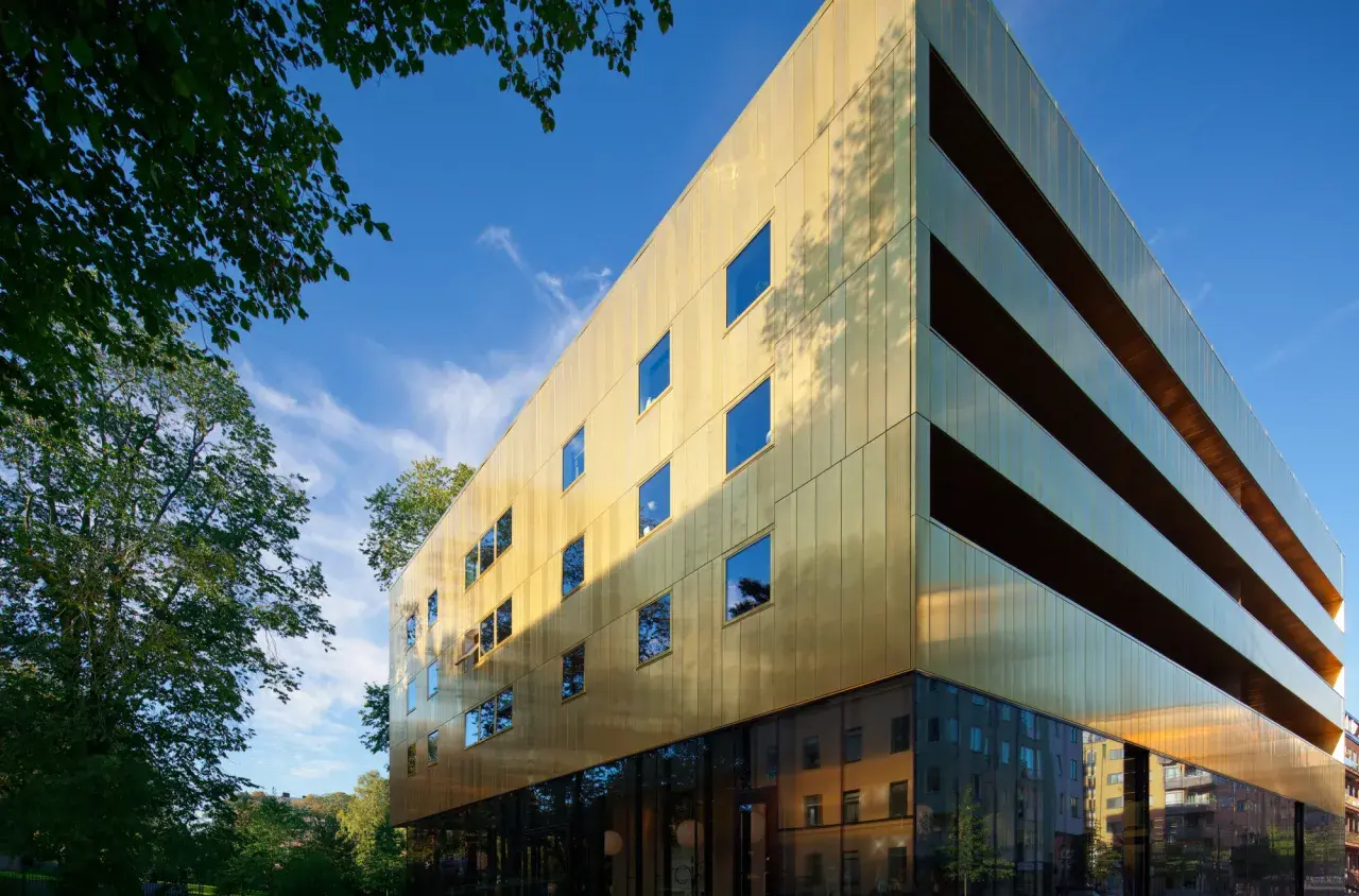 Sven-Harrys konstmuseum glänser i guld under en klarblå himmel. Gröna träd ramar in den moderna byggnaden.