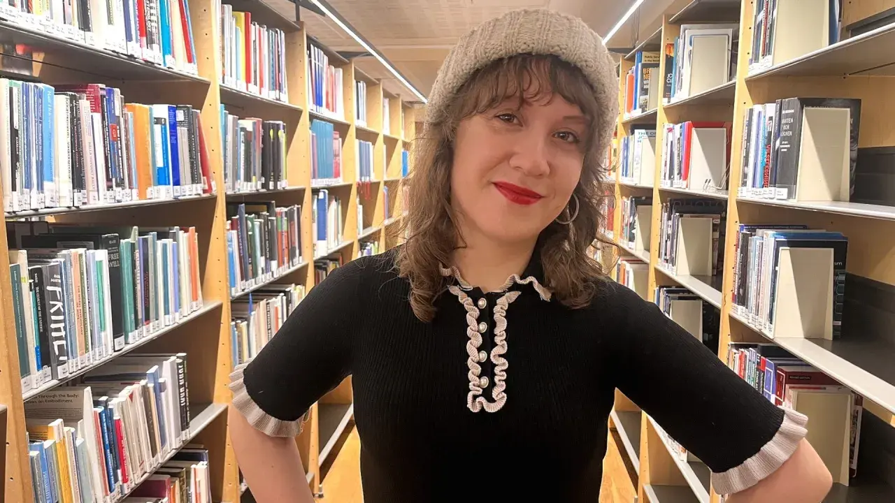 En kvinna med mössa står bland bokhyllor. Hon ler och undrar hur blir man bibliotekarie?
