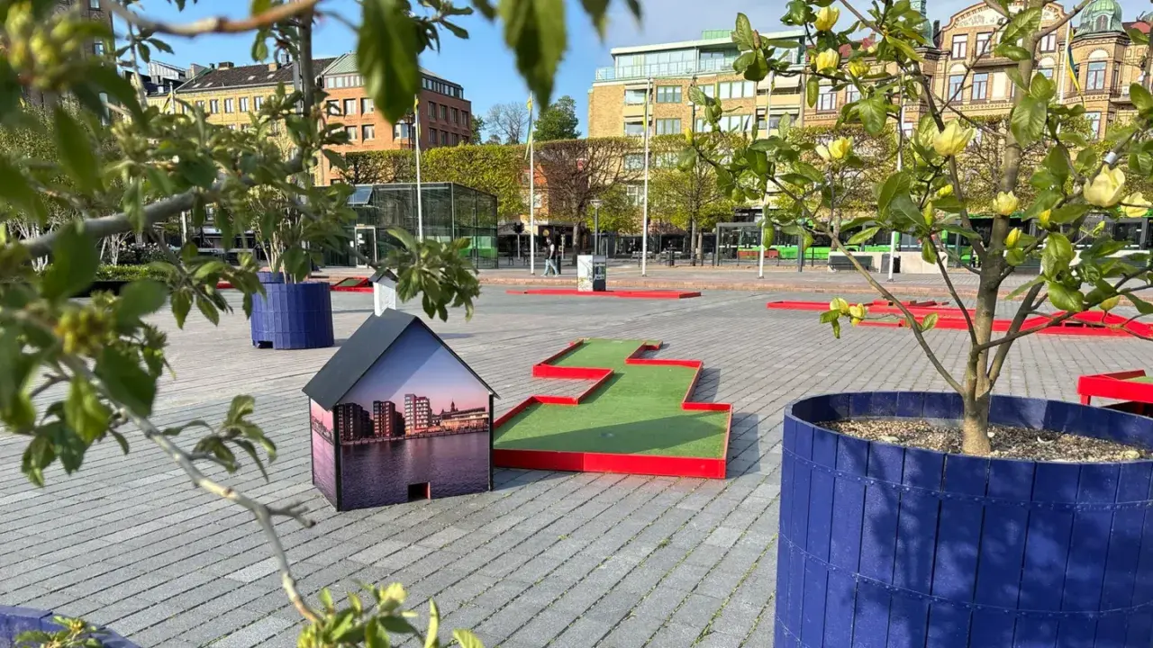 En minigolfbana i Helsingborg med en husmodell som visar stadens silhuett. Gröna träd och blå krukor ger liv åt platsen.