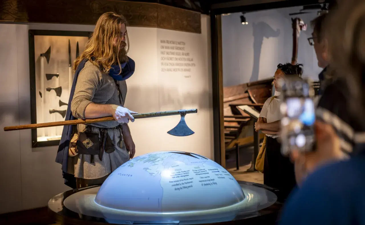 En guide i vikingakläder visar en yxa vid the viking museum. En jordglob med kartor och text projiceras.