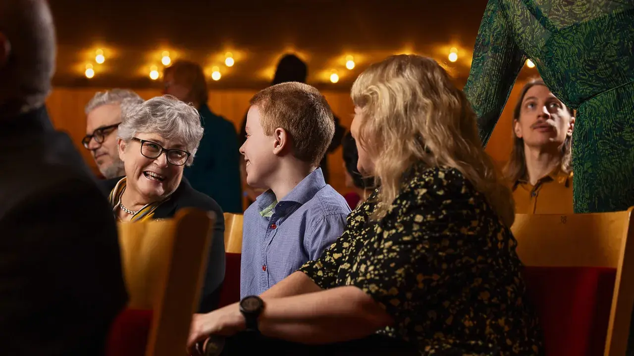 En familj njuter av en klassisk konsert. En mormor ler, ett barn tittar nyfiket framåt och en ung man i bakgrunden lyssnar uppmärksamt.