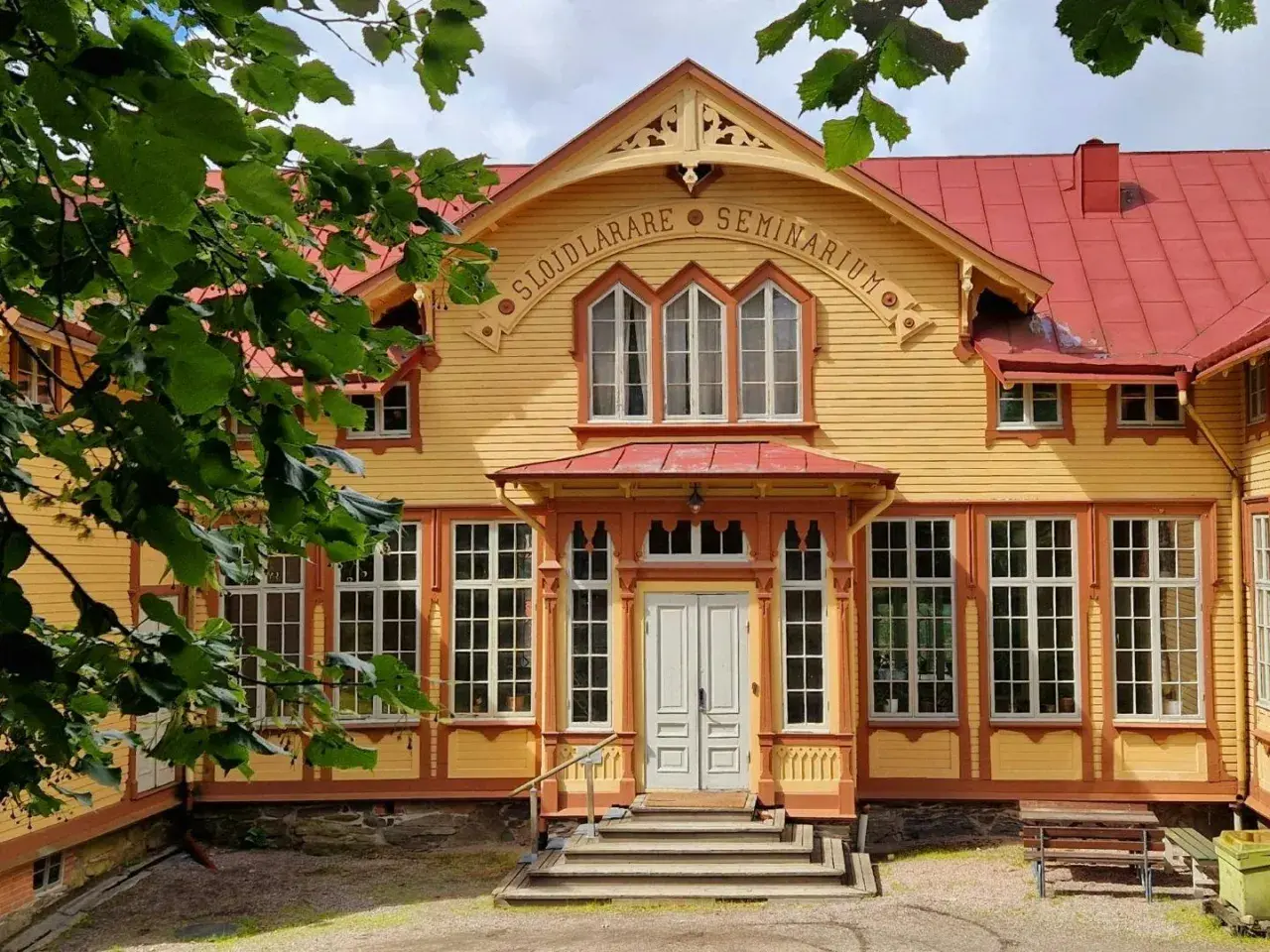 Gult trähus med röda detaljer, "Slöjdlärare Seminarium" text. Perfekt för en byggnadsvård kurs distans.