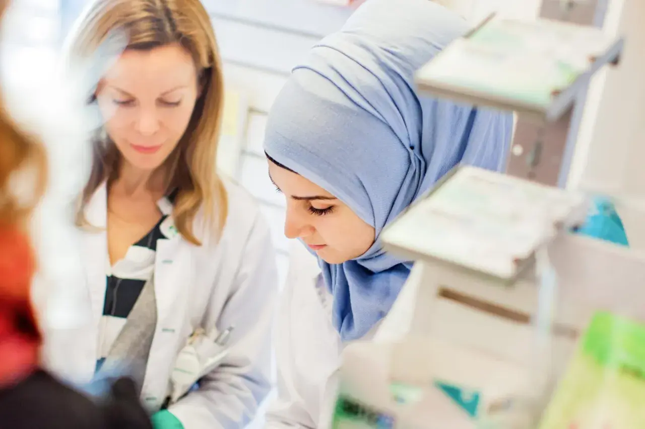 Två kvinnor i labbrockar, en med hijab, studerar något. De är på en arbetsplats som kan vara en del av en receptarie utbildning.
