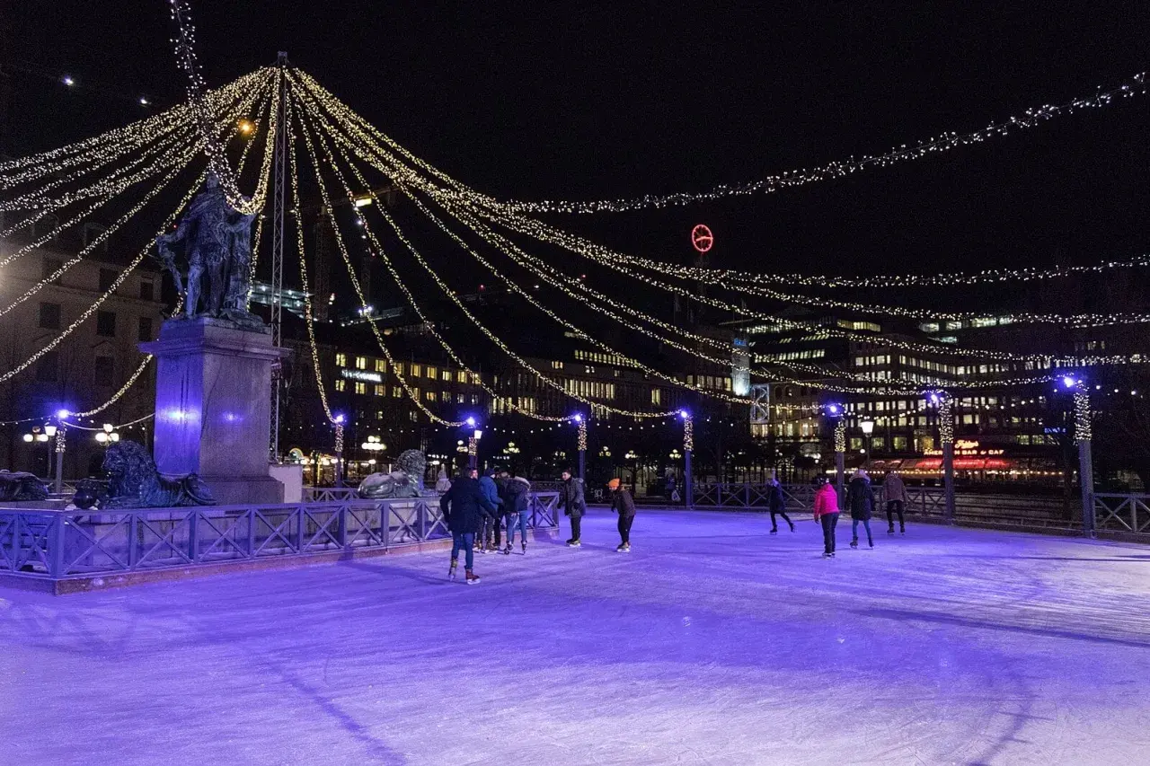 Människor åker skridskor på en upplyst isbana i Stockholm, omgivna av gnistrande ljusslingor och stadens silhuett.