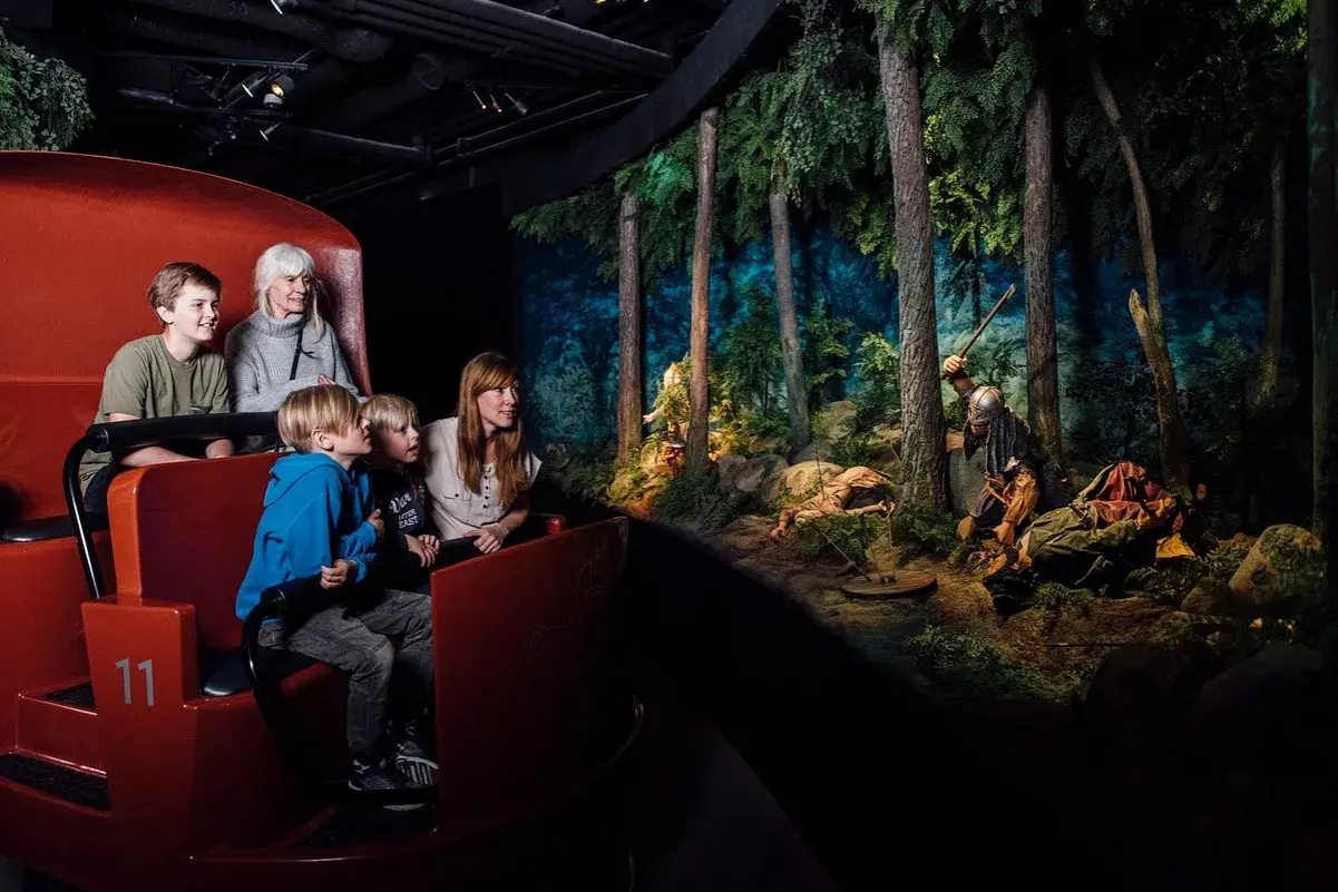 Familj i vagn på vikingamuseum, tittar på en dramatisk scen med vikingar i skogen. Bra recensioner för detta museum!