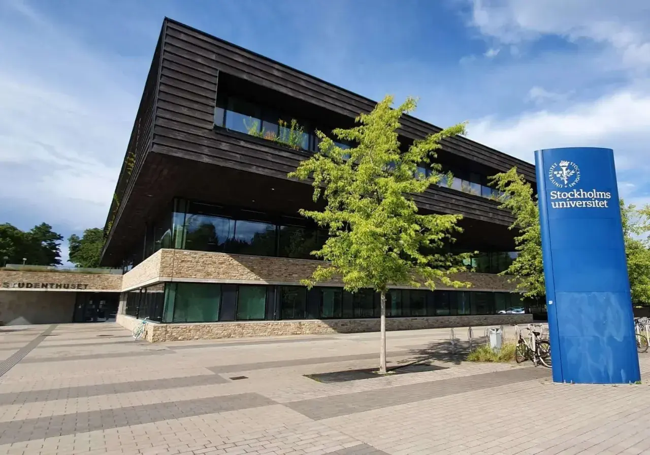 Modern byggnad med stenfasad och mörka träpaneler. En blå skylt med "Stockholms universitet" står bredvid. En del av universitet i Stockholm lista.