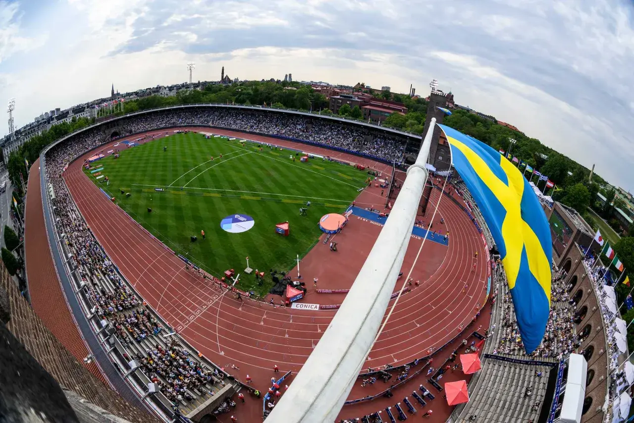 Stor svensk flagga vajar över en fullsatt arena i Stockholm. En friidrottstävling pågår på den röda löparbanan.