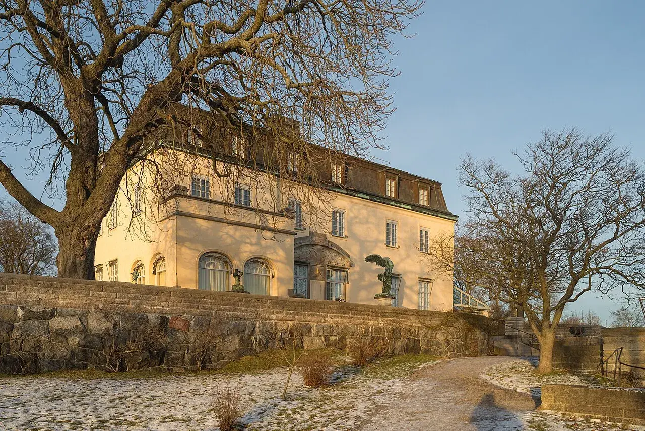 Prins Eugens Waldemarsudde i vinterskrud, med en stor ek och en staty framför den gula byggnaden.