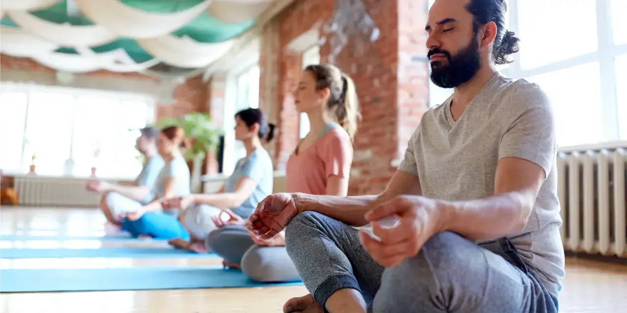 Människor med slutna ögon sitter i lotusställning på yogamattor. En grupp som deltar i en yoga utbildning distans.