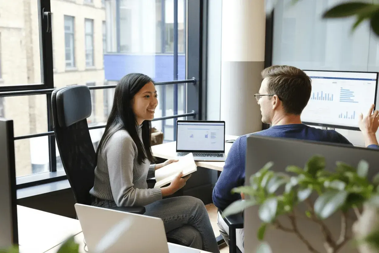 En leende kvinna håller en anteckningsbok medan hon pratar med en man som pekar på en datorskärm. De verkar diskutera affärsstrategier, kanske som en del av deras arbete som account manager.