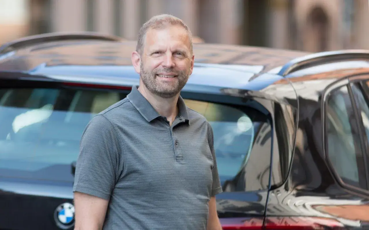 En glad man står bredvid en bil. Kanske funderar han på att leasa en begagnad bil för en bekväm resa.