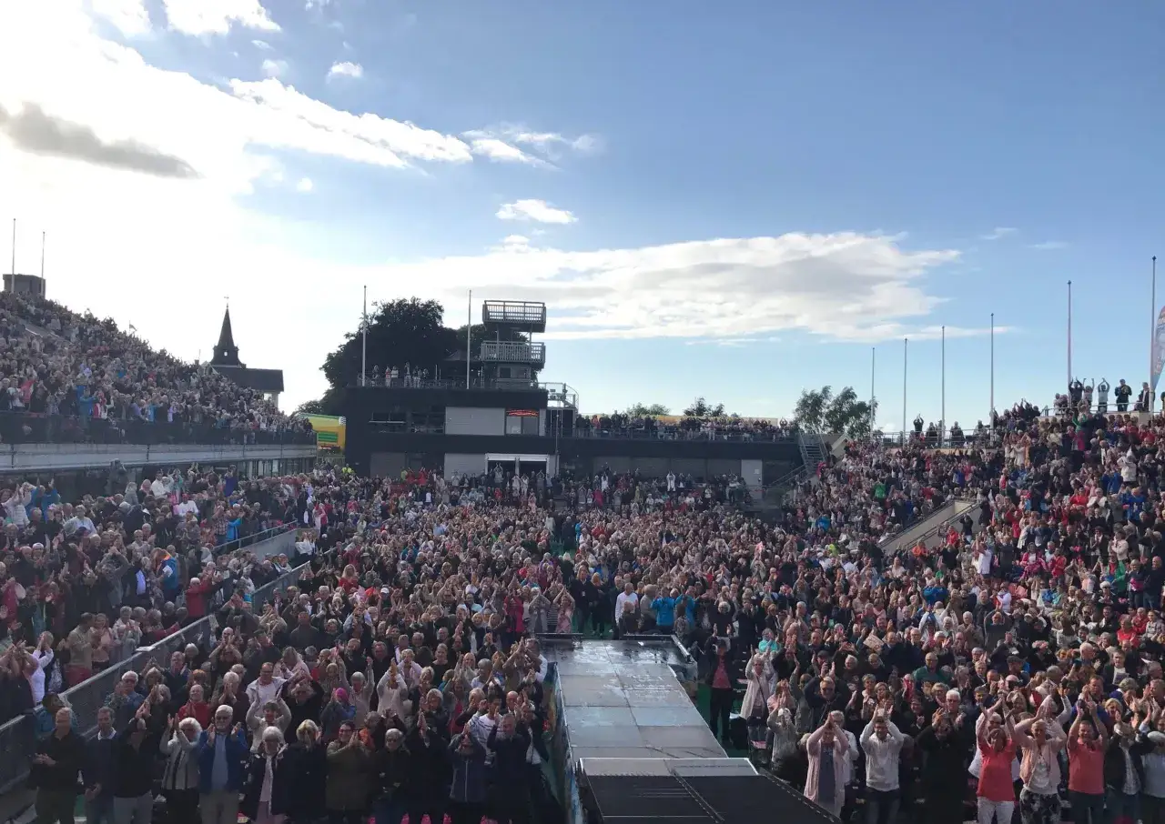 Stor publik applåderar vid Diggiloo i Båstad. Solen skiner på en molnig himmel över läktarna fyllda med glada åskådare.