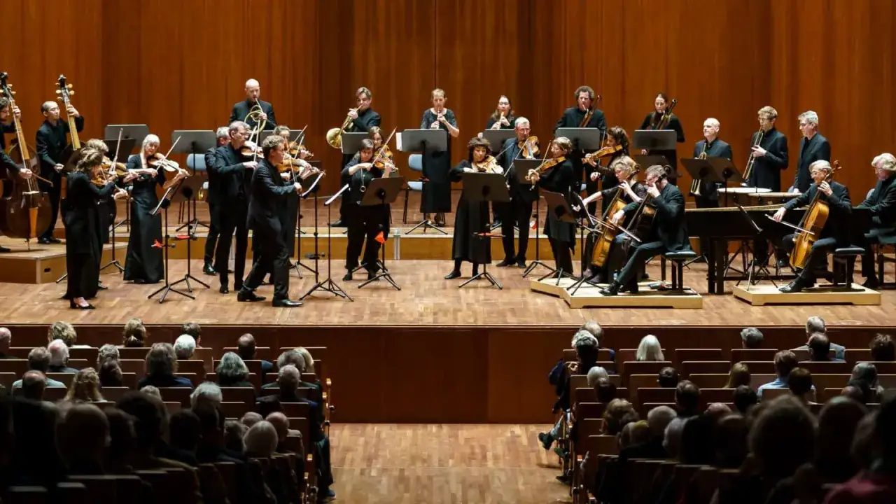 En orkester framför en klassisk konsert på en scen med träbakgrund. Publiken sitter i mörka stolar och lyssnar.