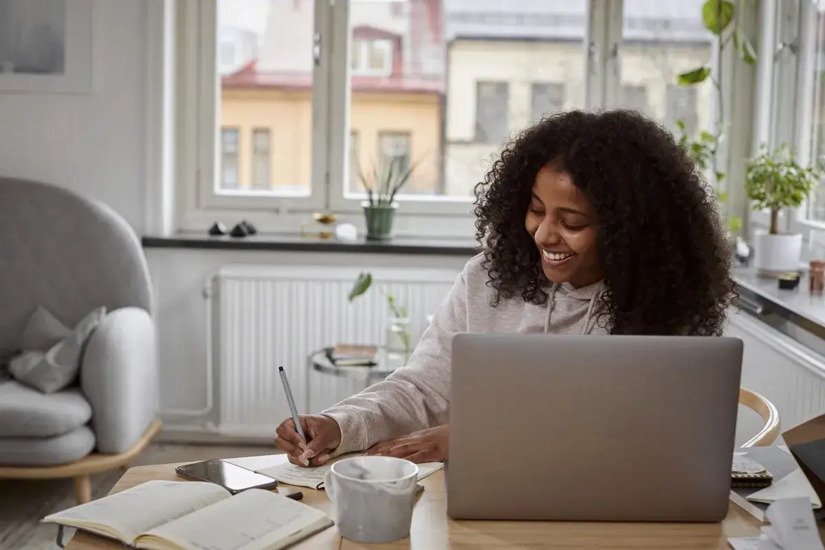 Glad kvinna pluggar fristående kurser distans vid sin laptop. Hon skriver anteckningar i en bok.