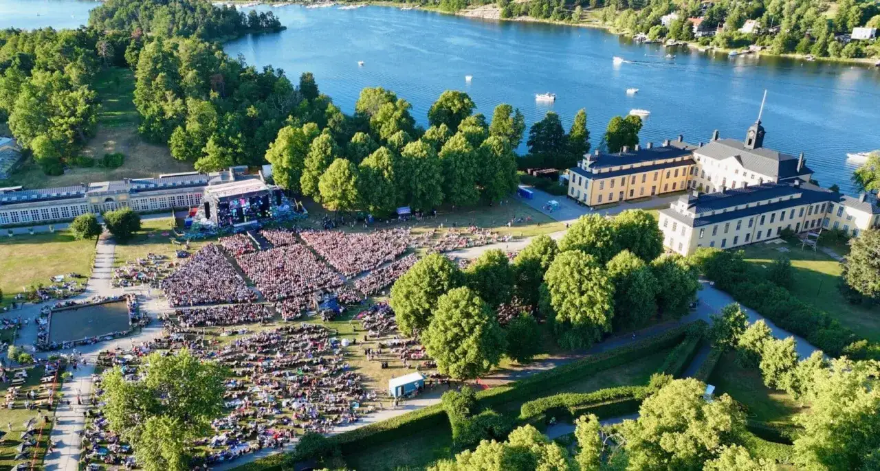 Stor publik njuter av en gratis konsert i Stockholm vid vattnet, med en imponerande scen och historiska byggnader i bakgrunden.