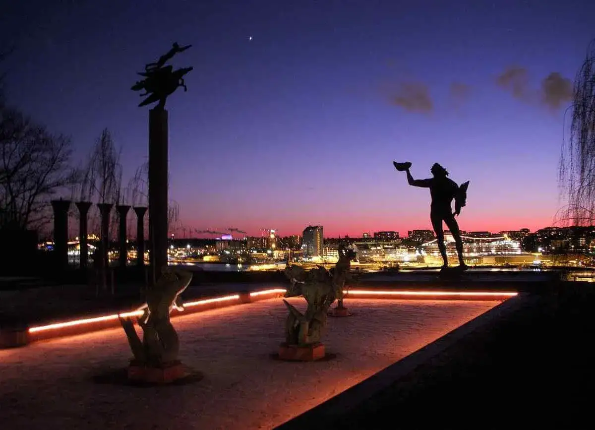 Skymning över Stockholm med konstverk som en del av ett upplevelsemuseum. Silhuetter av statyer mot en färgsprakande himmel och stadens ljus.