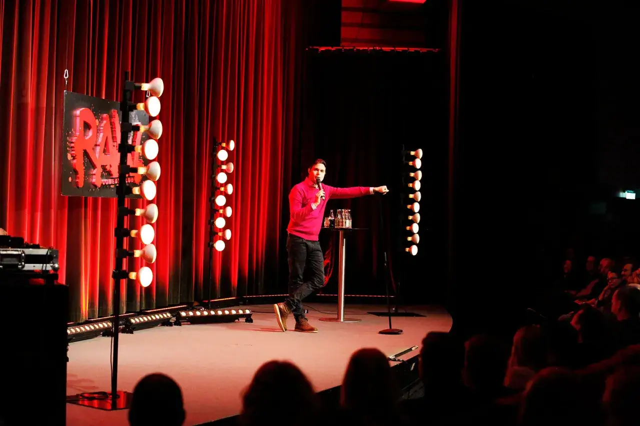 En komiker på scenen, redo för standup Sverige. Röda draperier och lampor skapar en intim atmosfär.