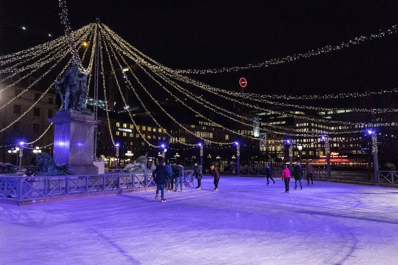 People ice skating at night on a rink illuminated by purple lights and decorated with strings of golden fairy lights.