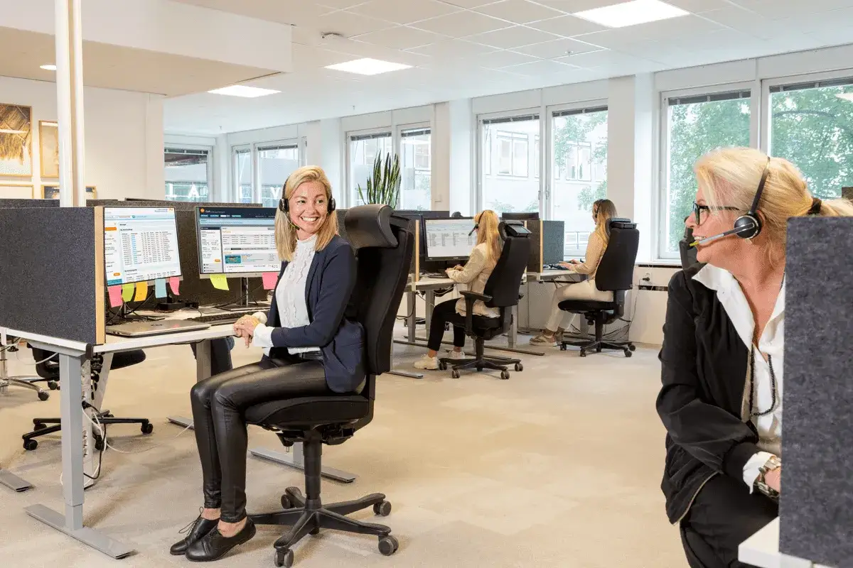 Glad account manager ler vid sitt skrivbord med headset. Kollegor arbetar vid sina datorer i bakgrunden.