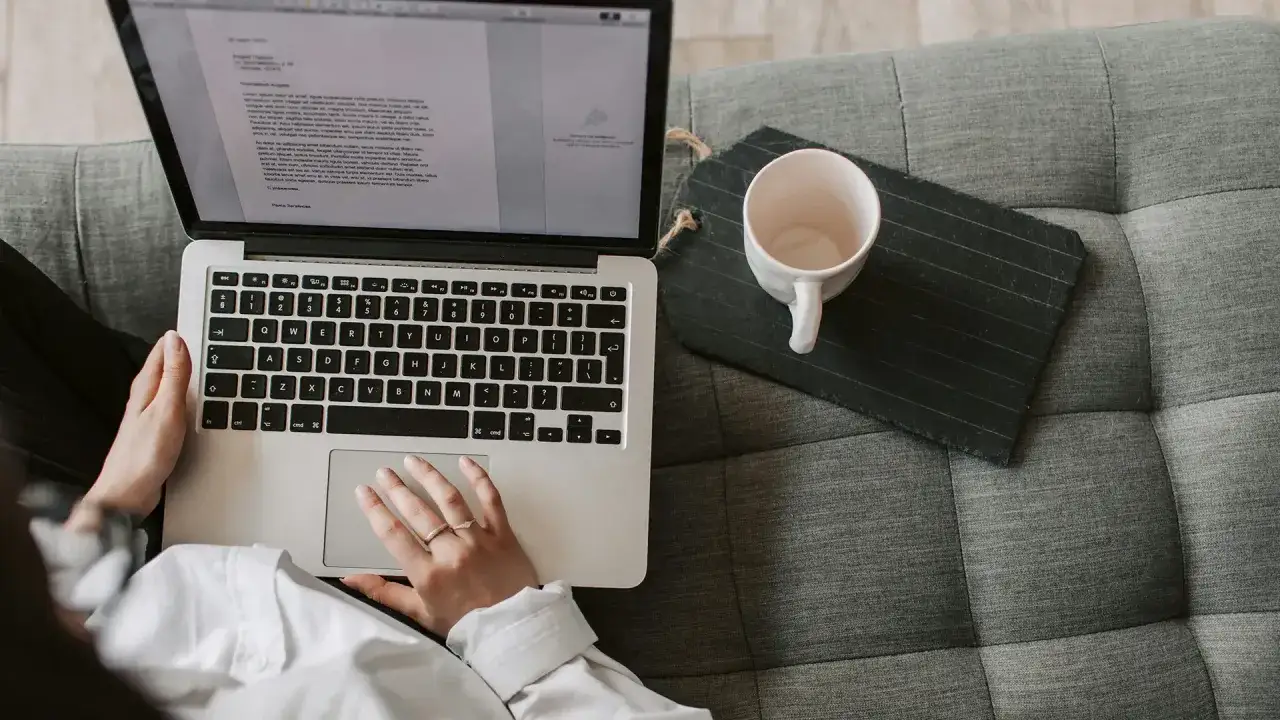 Person studerar konstvetenskap distans på en laptop i soffan, med en kaffekopp bredvid.