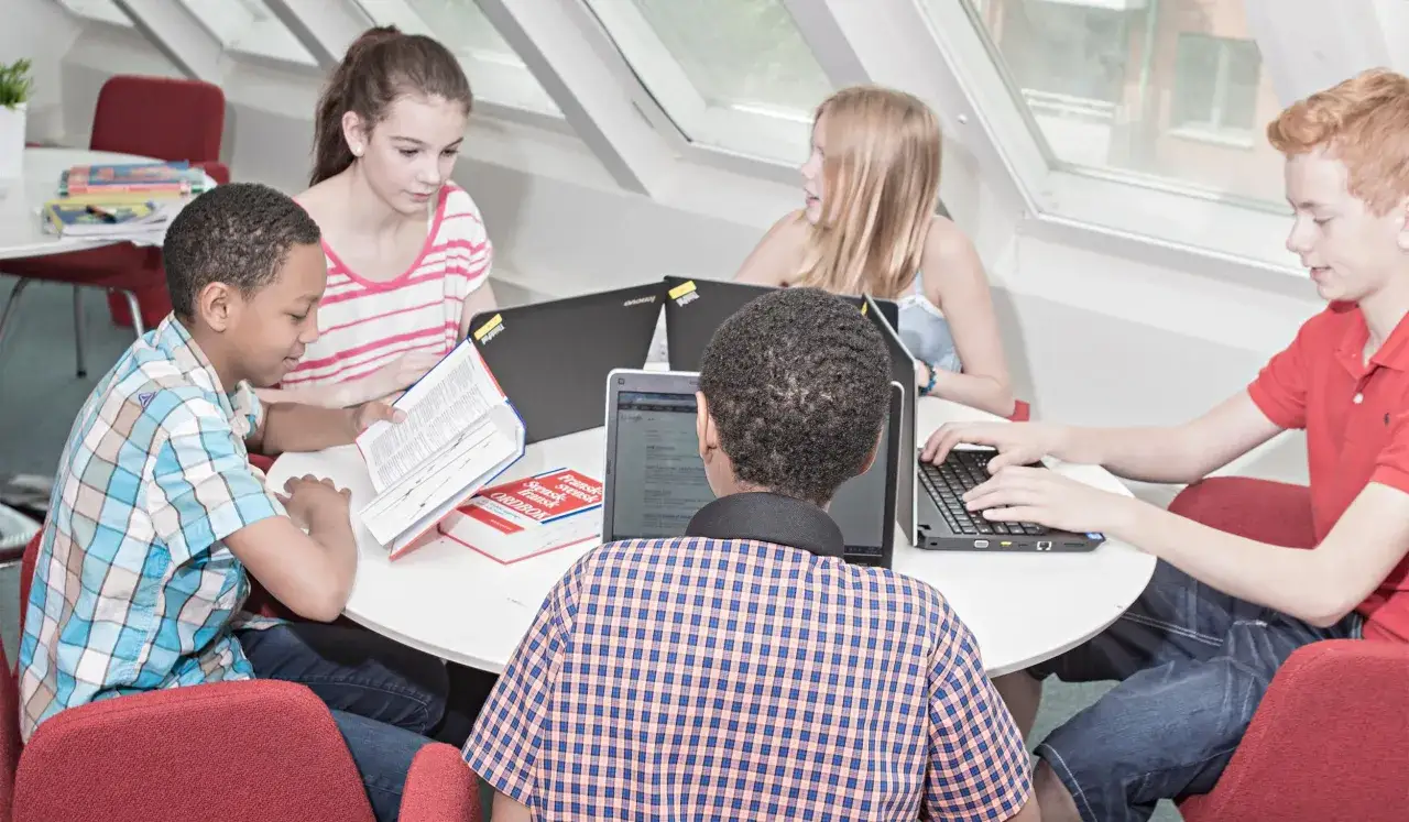 Barn samarbetar kring en academy online-uppgift med böcker och laptops.